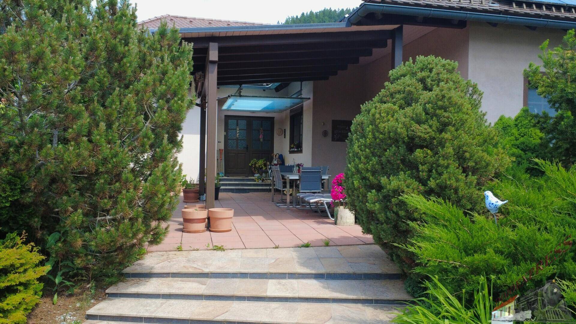 Überdachte Terrasse mit Gartenmöbeln, umgeben von üppigem Grün und Sträuchern.