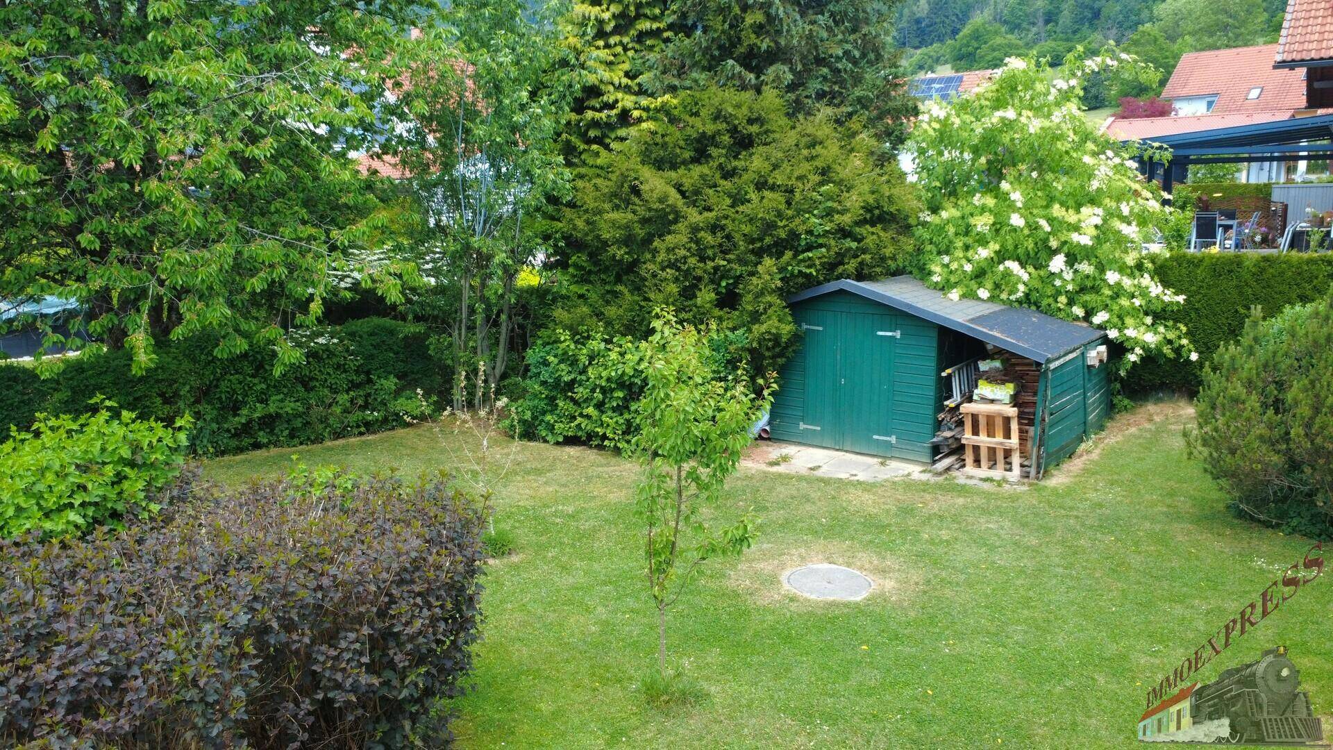 Grüner Gartenbereich mit einem kleinen Geräteschuppen und verschiedenen Pflanzen und Sträuchern.