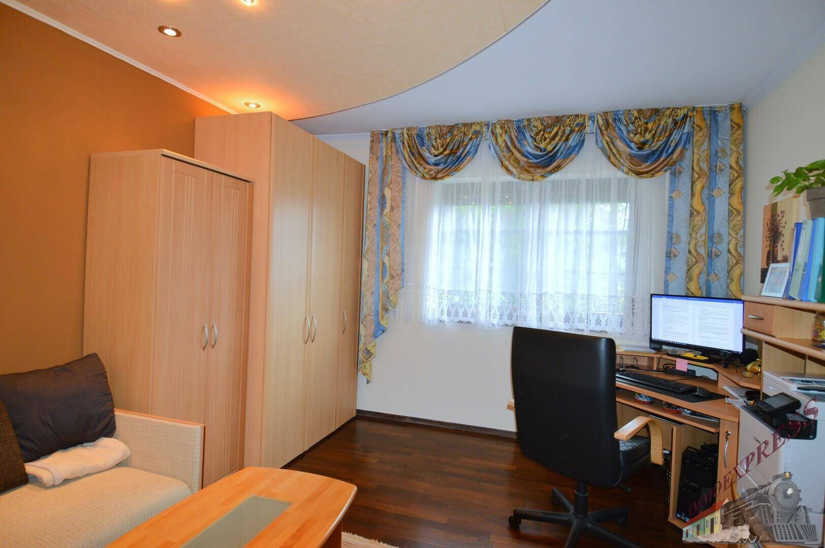 Schlafzimmer mit Schreibtisch, Kleiderschrank und Fenster mit Vorhängen.