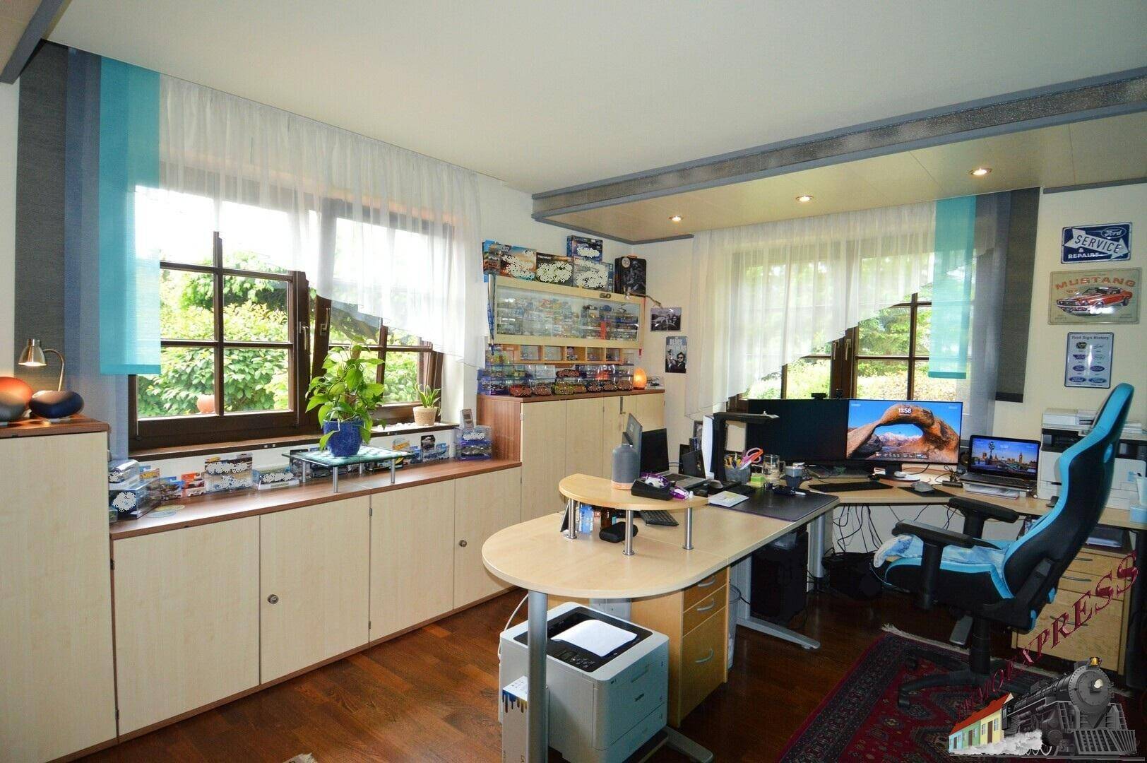 Helles Arbeitszimmer mit großem Schreibtisch, Bürostuhl und Fenstern mit Blick ins Grüne.