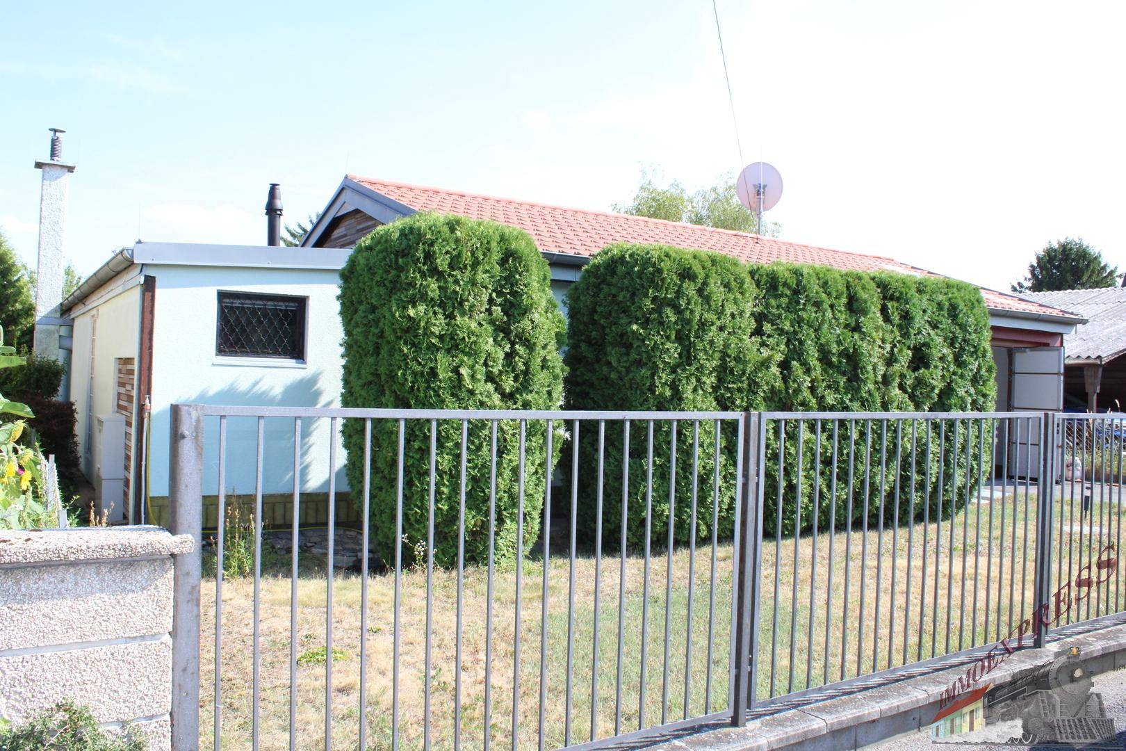 Seitenansicht des Hauses mit einem Zaun und einer Reihe von hohen Hecken unter blauem Himmel.