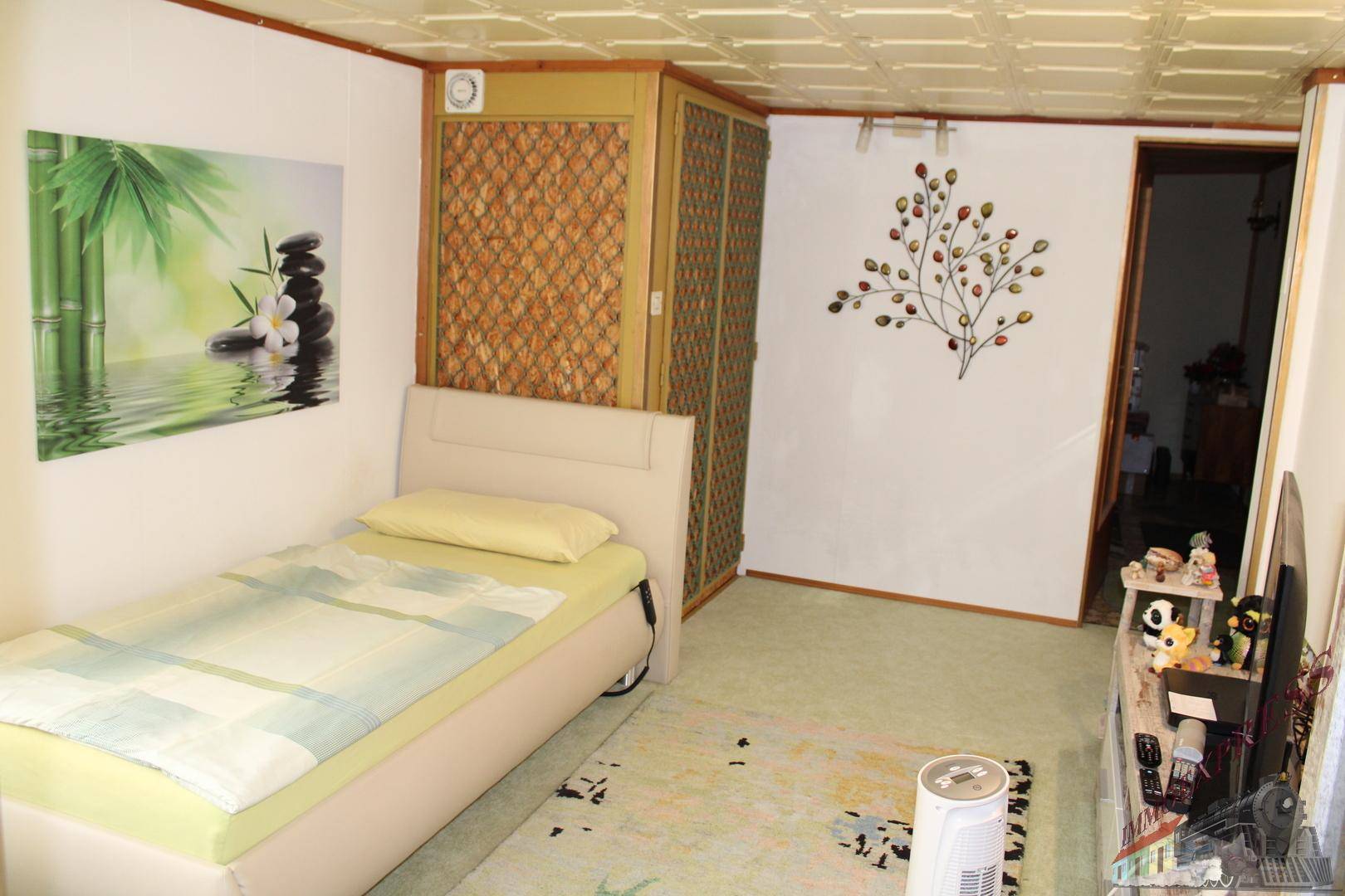 Schlafzimmer mit Einzelbett, Teppichboden und einer dekorativen Deckenverkleidung, hell gestaltet.