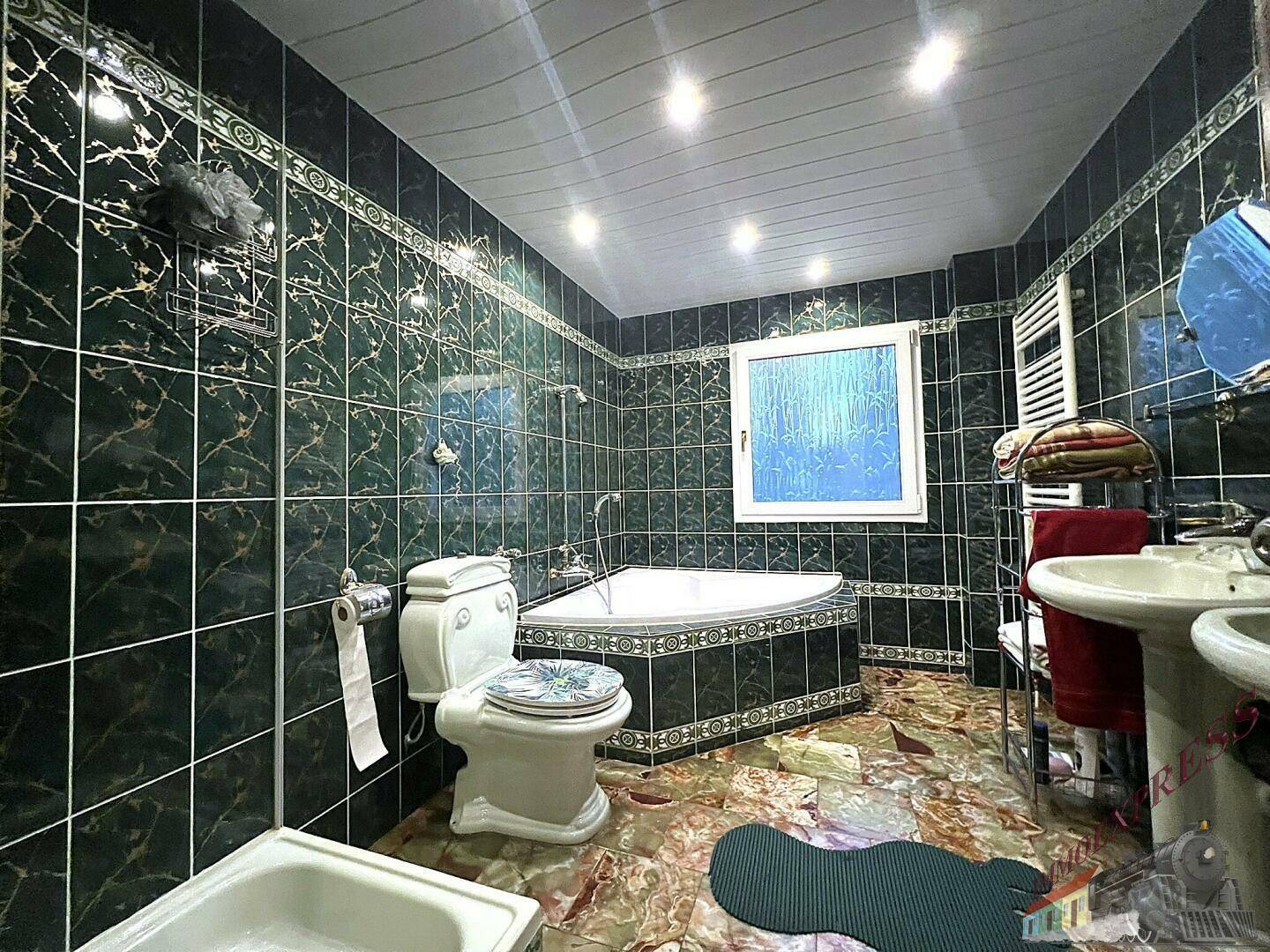 Badezimmer mit dunklen Fliesen, Eckbadewanne und einem Fenster für natürliches Licht.