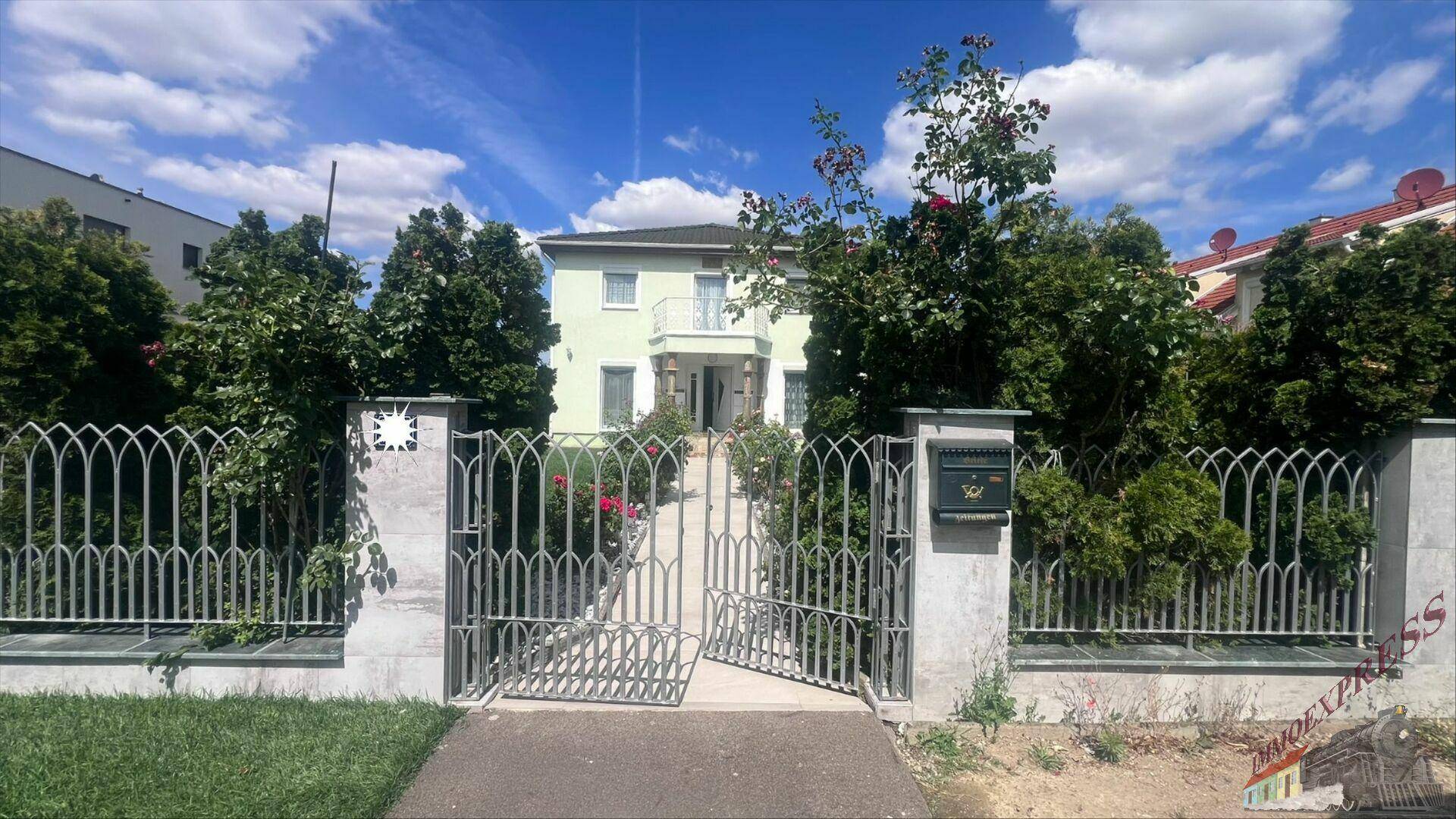 Das Haus ist von einem dekorativen Metallzaun umgeben, mit üppigem Grün im Vorgarten und Bäumen.
