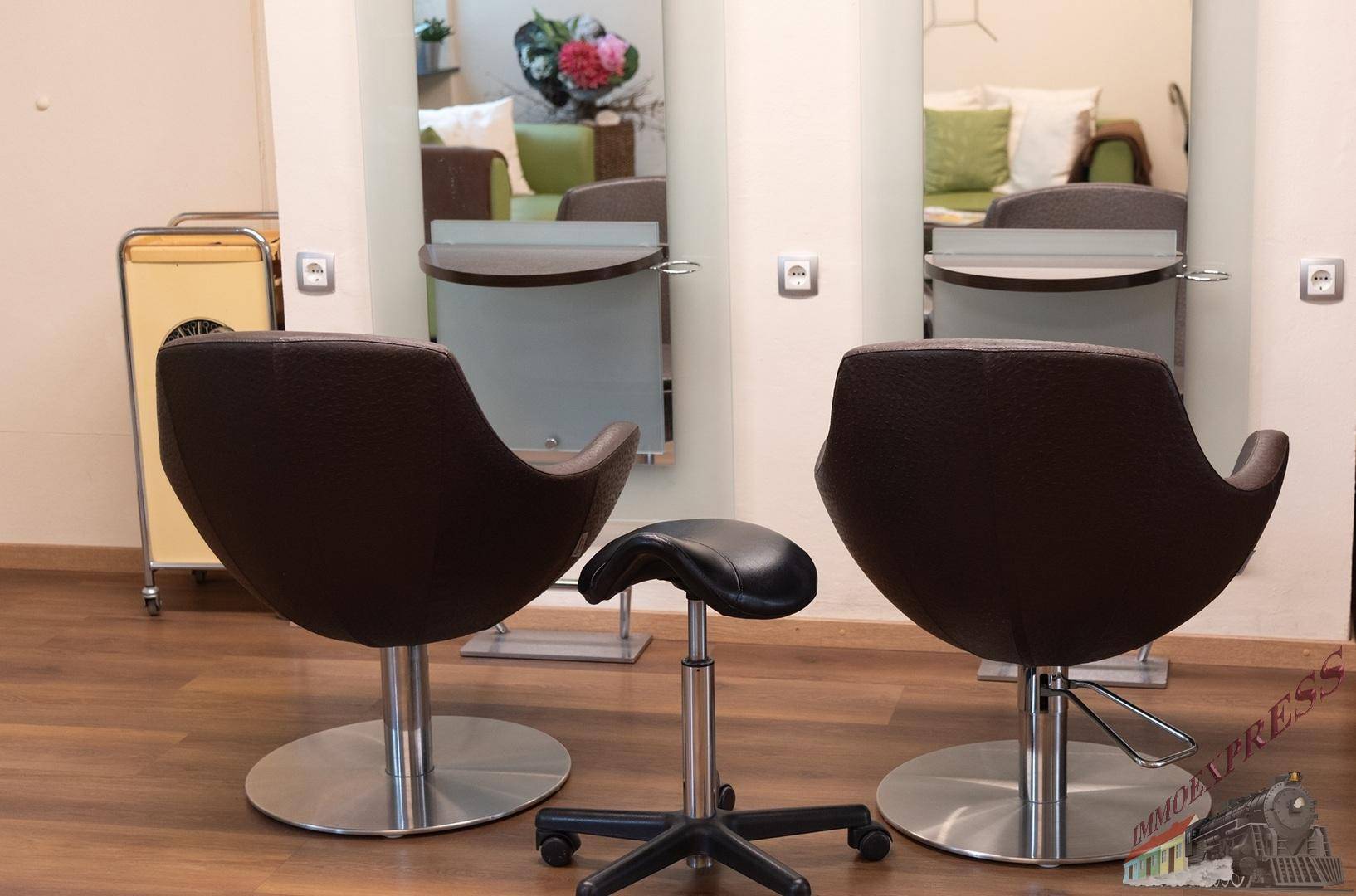 Zwei Styling-Stühle und ein Hocker vor Spiegeln, die den Wartebereich reflektieren.