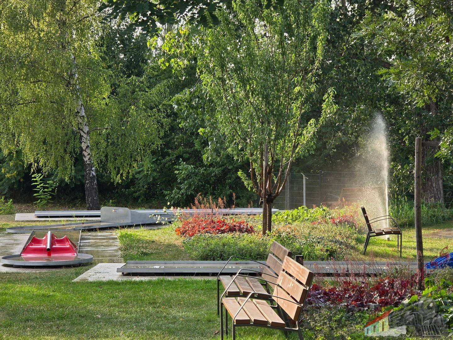 Detailansicht einer Minigolfbahn mit Wasserspiel, umgeben von Blumenbeeten und einer Holzbank.
