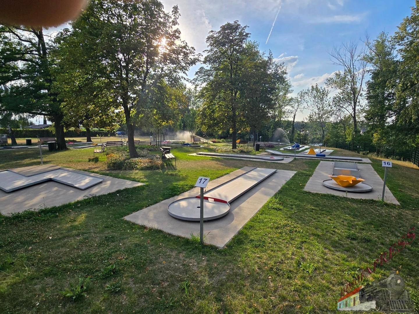 Sonnige Minigolfanlage mit mehreren Bahnen, umgeben von Bäumen und bewässertem Rasen.