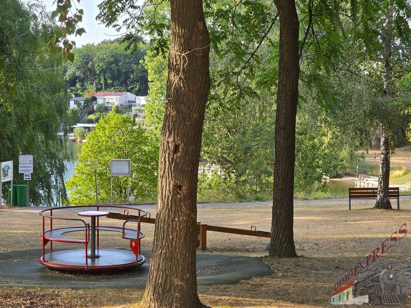 Spielplatz mit Karussell und Wippe, im Hintergrund ist der See und weitere Bäume sichtbar.