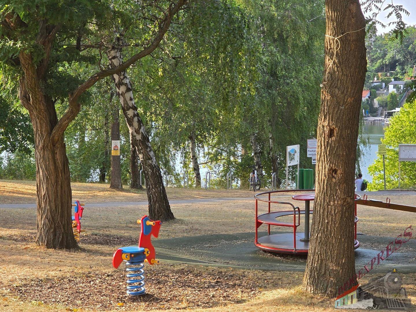 Spielplatz mit Karussell und Federwippe, umgeben von Bäumen und mit Blick auf den See.
