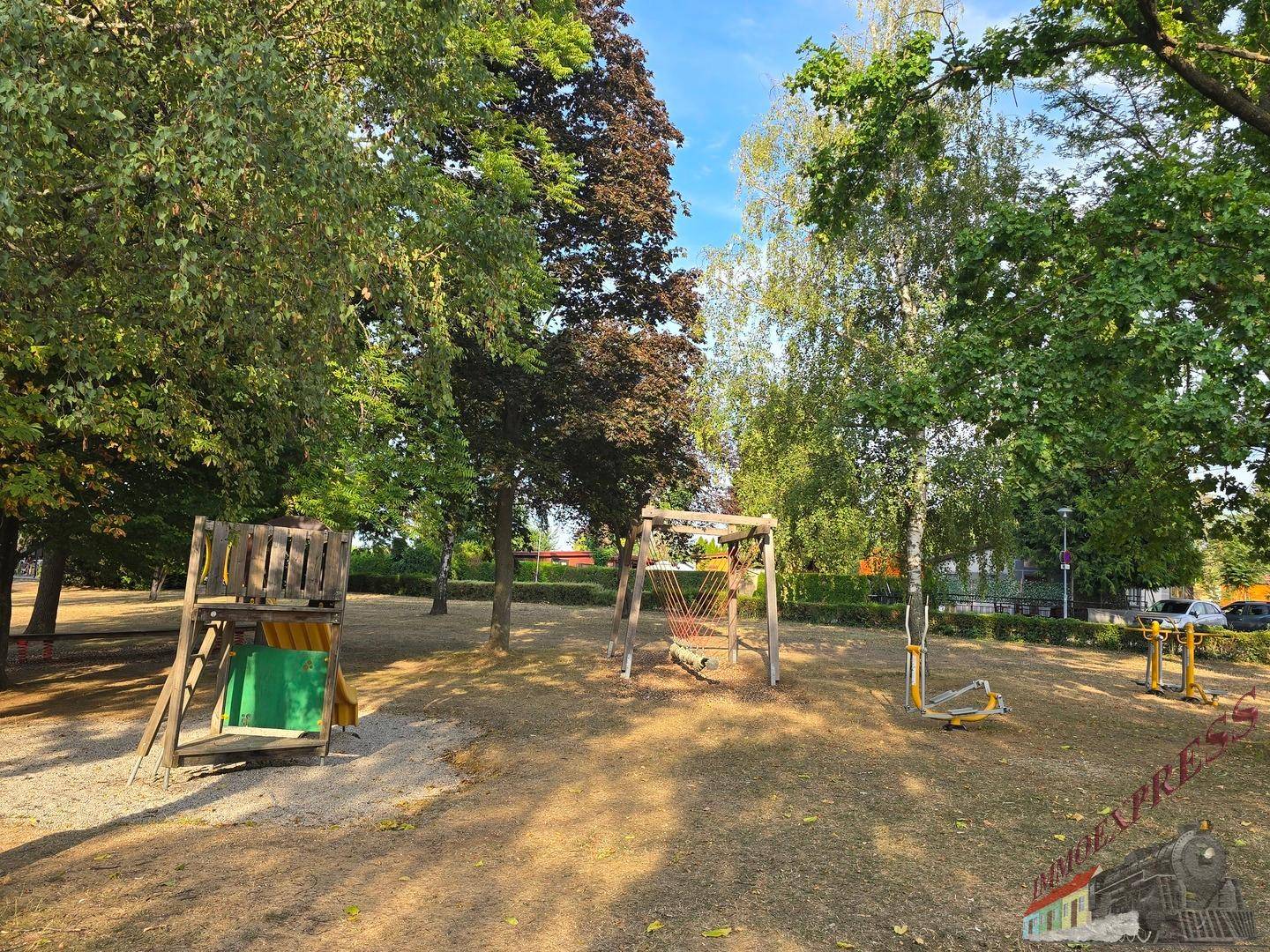 Spielplatz mit Holzspielgeräten, Schaukeln und Fitnessgeräten im Freien, umgeben von Bäumen.