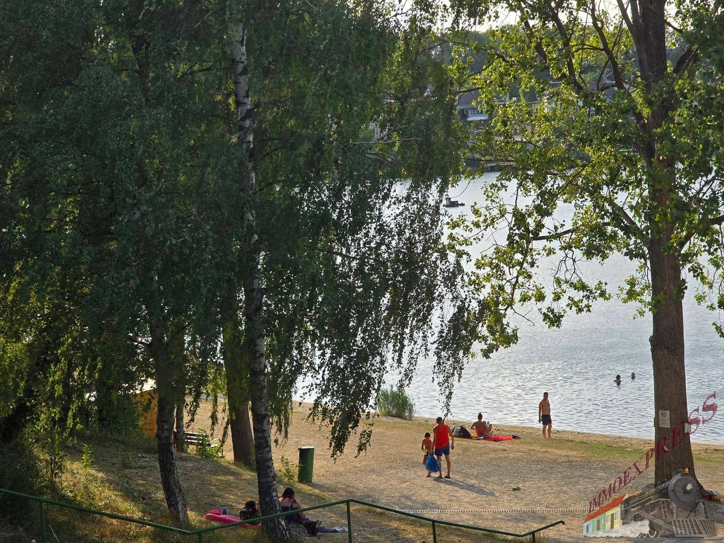 Sandiger Badestrand am Seeufer mit Badegästen, umgeben von schattenspendenden Bäumen.