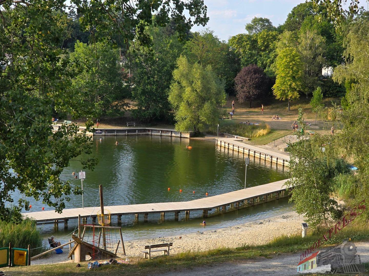 Seeufer mit Badestrand, Holzstegen und Spielbereich, umgeben von Bäumen und Grünflächen.