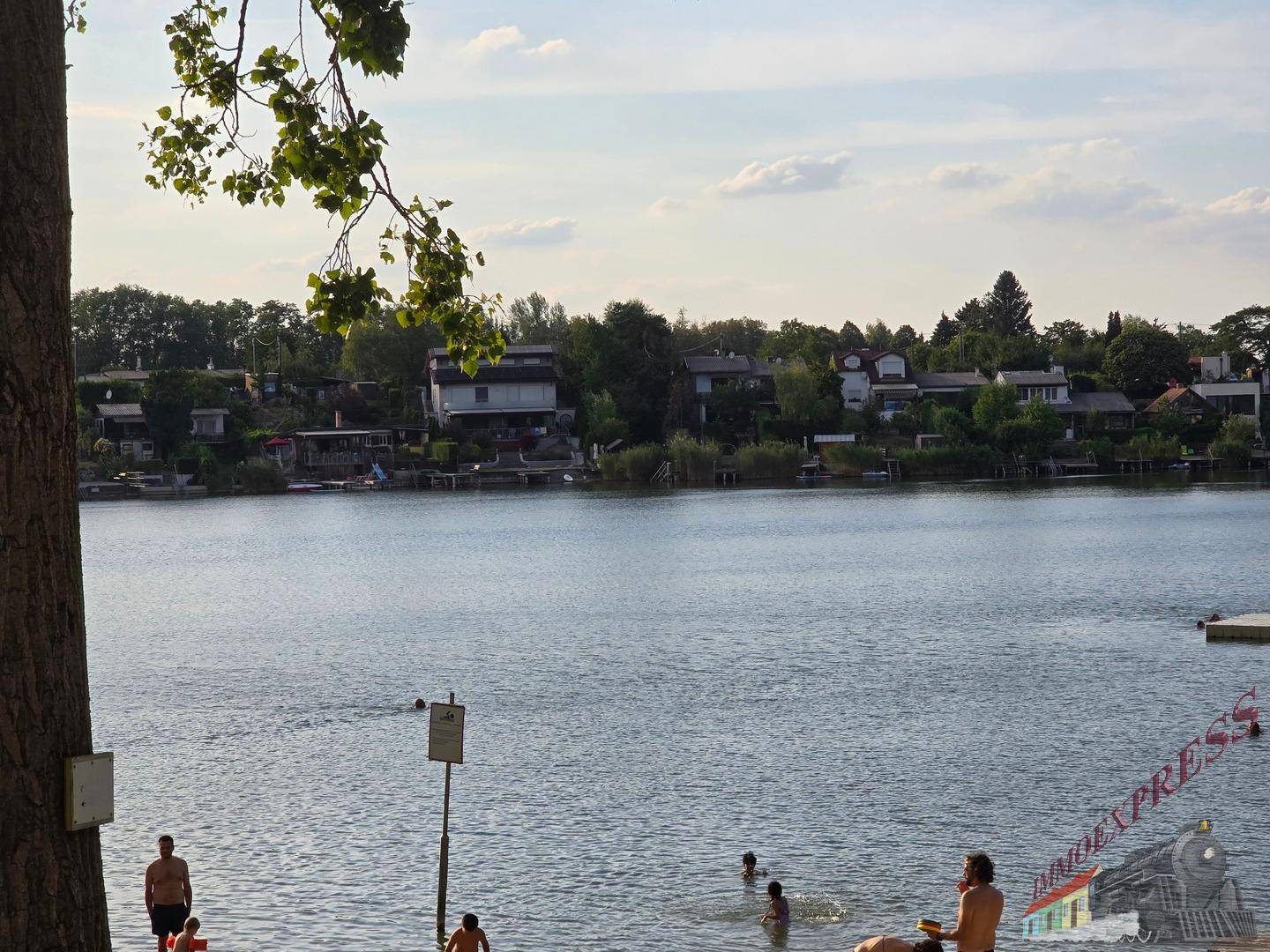 Weitläufiger Seeblick mit Häusern am gegenüberliegenden Ufer und Badegästen im Vordergrund.