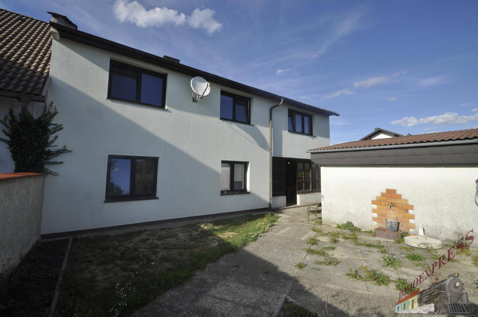 Rückansicht des Hauses mit weißer Fassade, dunklen Fenstern und gepflastertem Bereich.