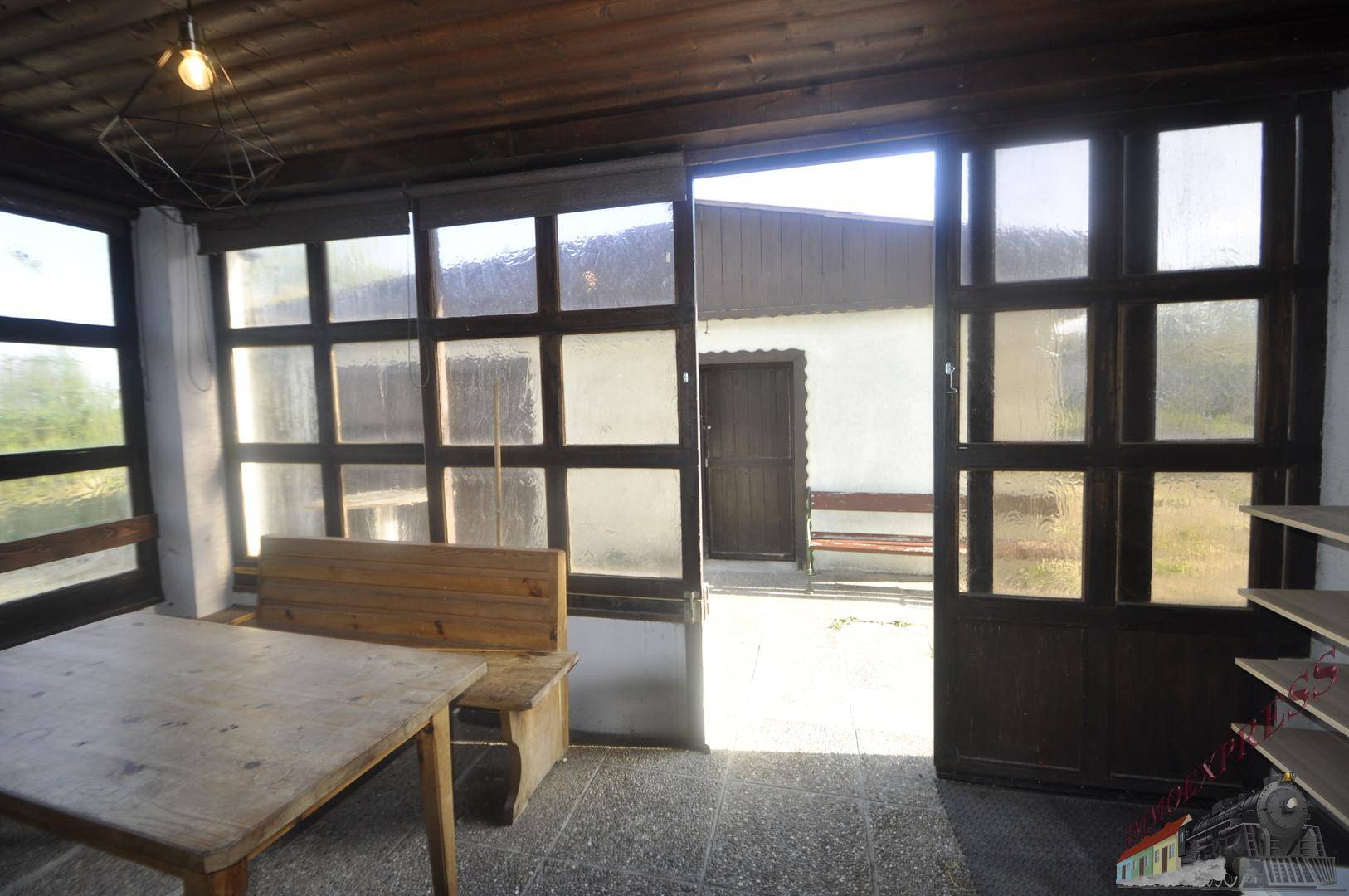 Heller Wintergarten mit großen Glasfenstern, Holztisch und Bank, der zu einem Außenbereich führt.
