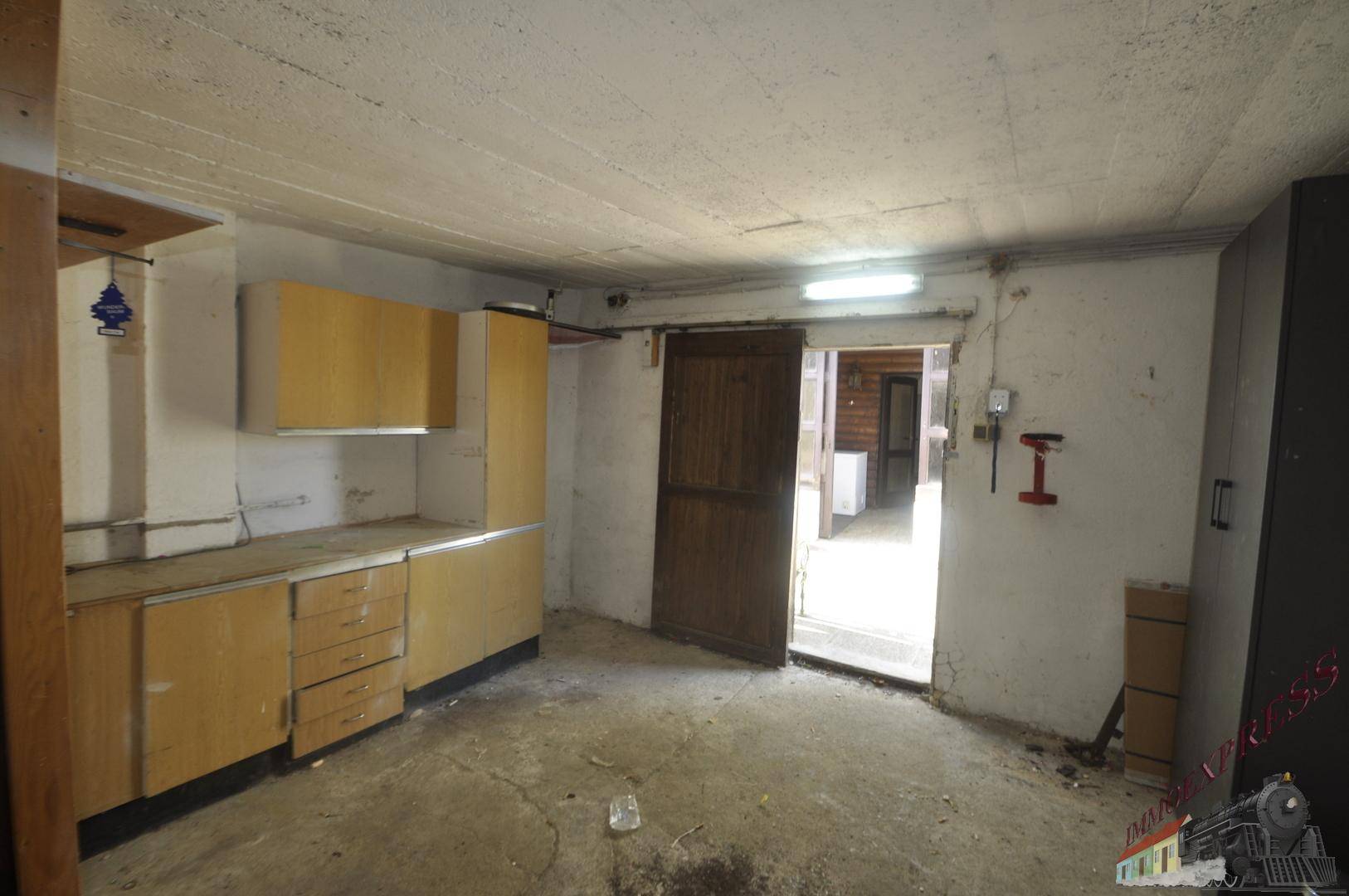 Kellerraum mit älteren Küchenschränken, Betonboden und einer Tür, die ins Freie führt.