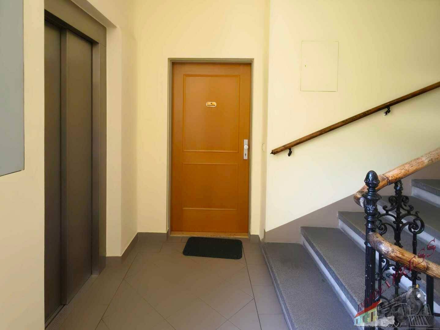 Treppenabsatz mit Aufzug, Wohnungstür und Treppe mit schmiedeeisernem Geländer.
