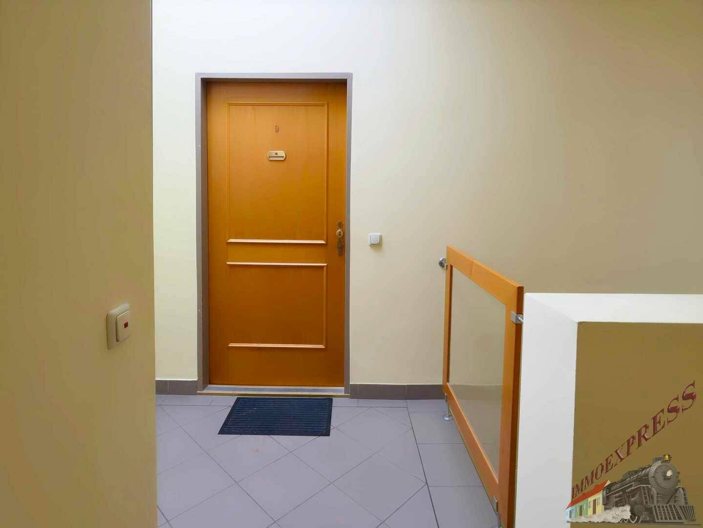 Heller Flur mit einer Wohnungstür, Fliesenboden und einem modernen Geländer.