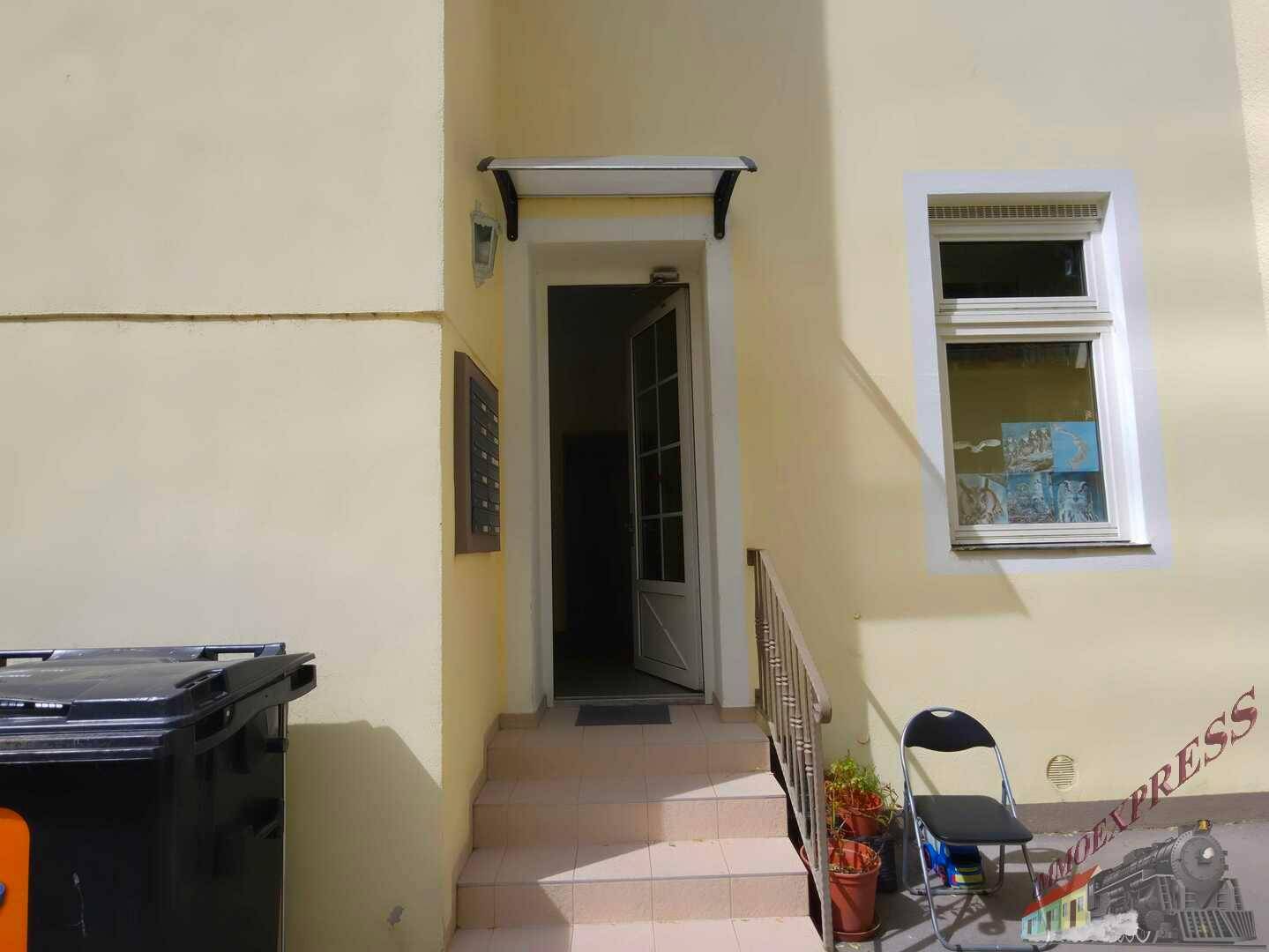 Eingangsbereich eines Gebäudes mit Treppe, Vordach und Mülltonnen im Freien.