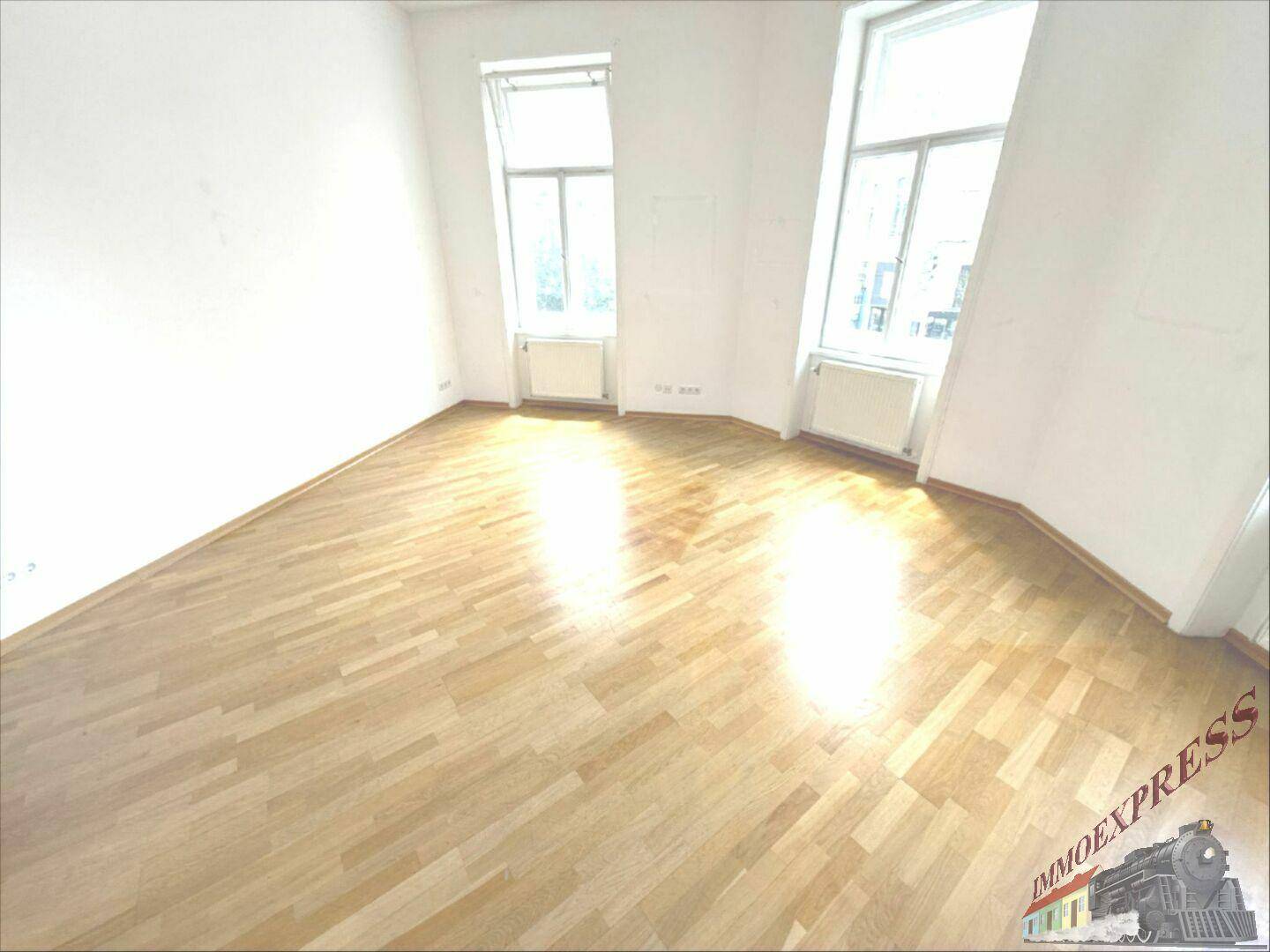 Leerer Raum mit hellem Holzparkettboden und zwei hohen Fenstern, die viel Tageslicht hereinlassen.