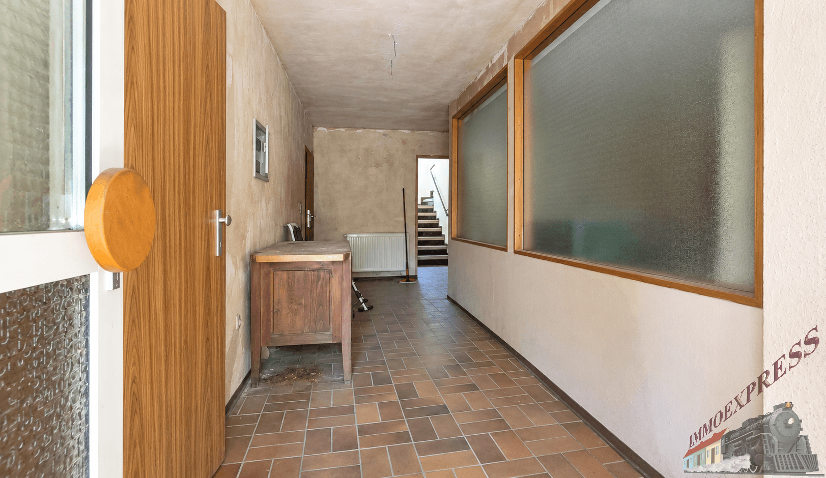 Geräumiger Flur mit Terrakotta-Fliesenboden, Holztür und einer Holzkonsole, Treppe im Hintergrund.
