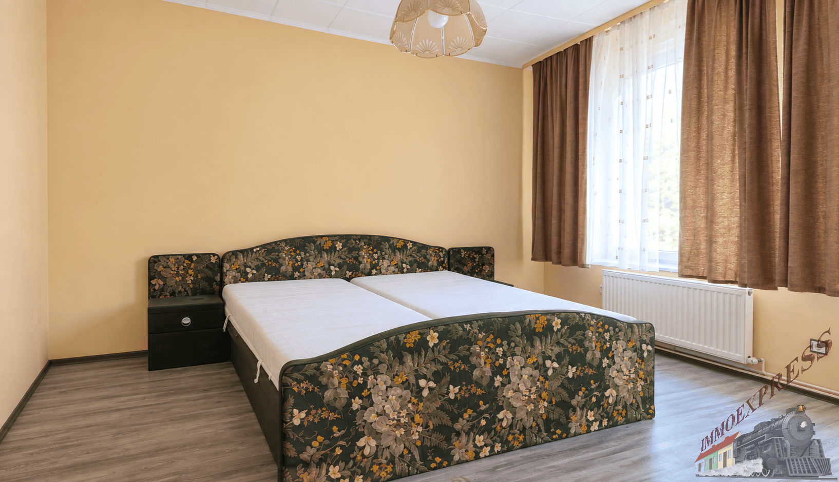 Schlafzimmer mit Laminatboden, einem Doppelbett mit Blumenmuster und Fenster mit braunen Vorhängen.