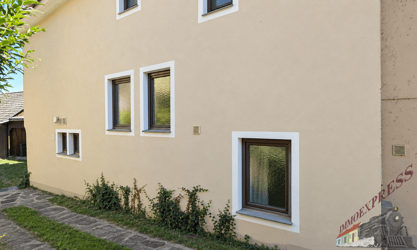 Seitenfassade des Hauses mit mehreren Fenstern und leichter Begrünung am Sockel.