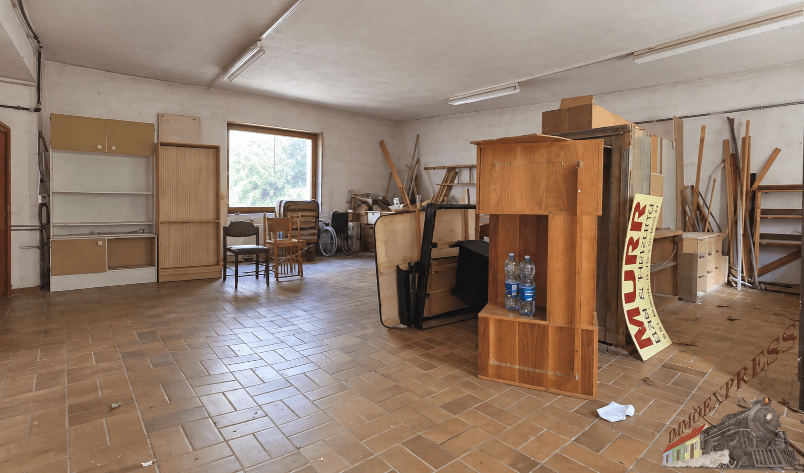Großer leerer Raum mit Fliesenboden und Fenster, genutzt zur Lagerung von Möbeln und Materialien.