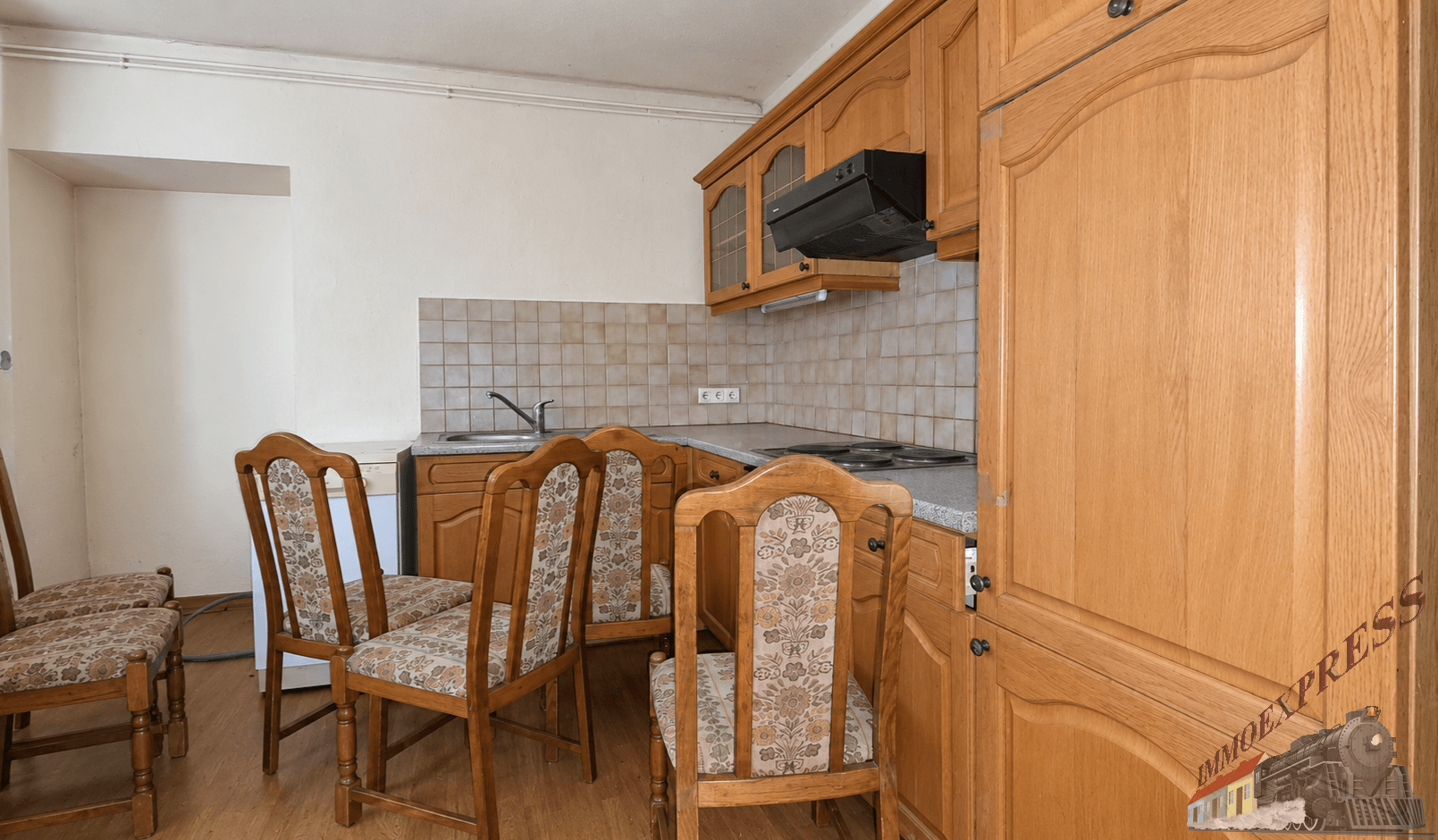 Einfache Küche mit Holzschränken, Fliesenrückwand und traditionellen Esszimmerstühlen.