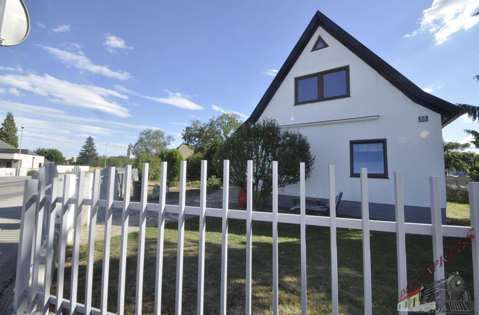 Einfamilienhaus mit weißer Fassade und Satteldach, umgeben von einem weißen Zaun und Grünflächen.