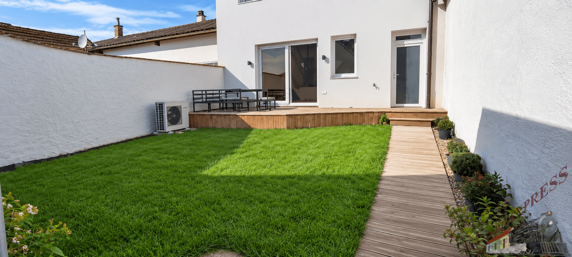 Sonniger Gartenbereich mit frischem Rasen, Holzterrasse und weißer Hausfassade.