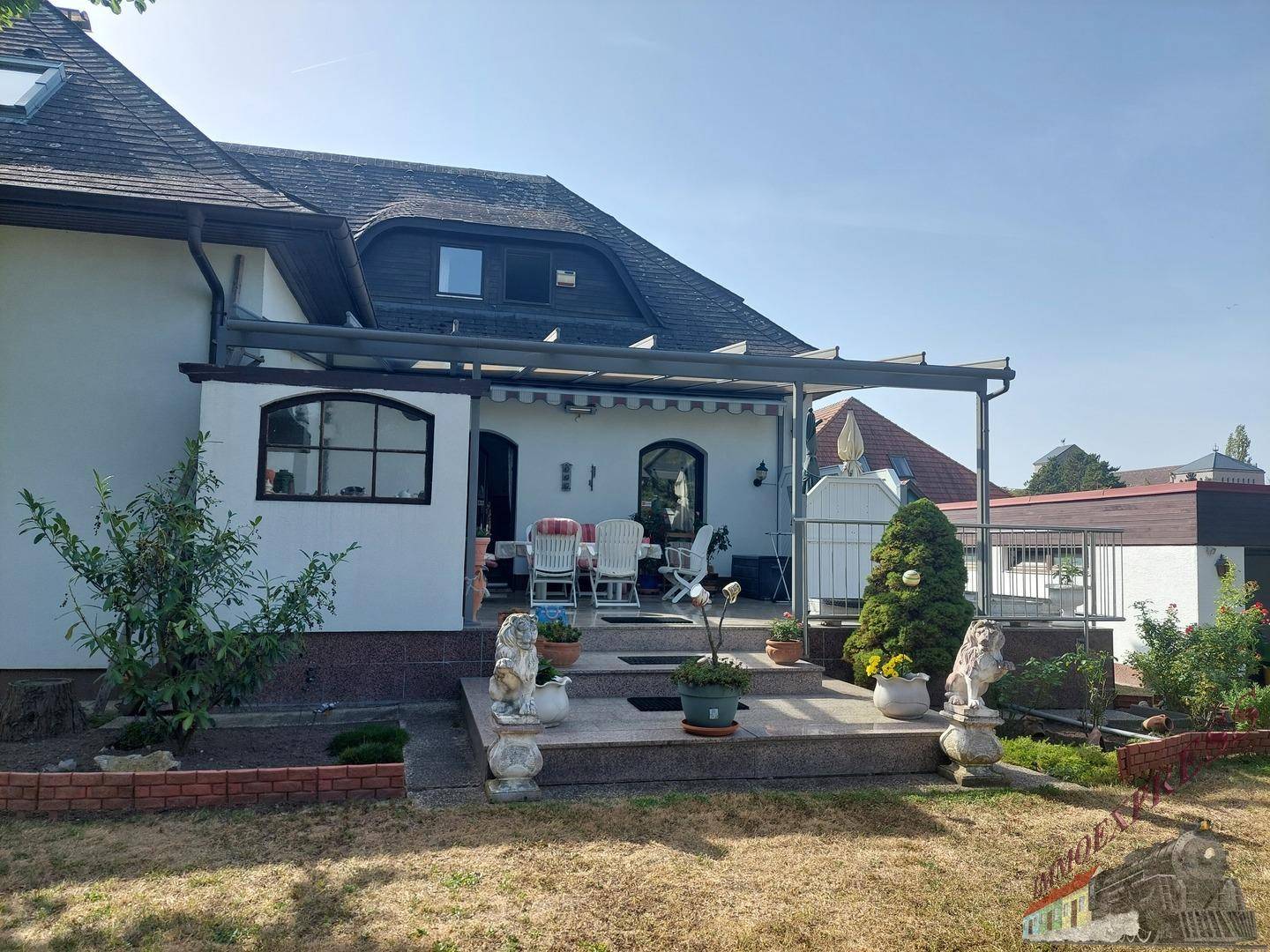 Rückansicht des Hauses mit großer Terrasse und gepflegtem Garten unter blauem Himmel.