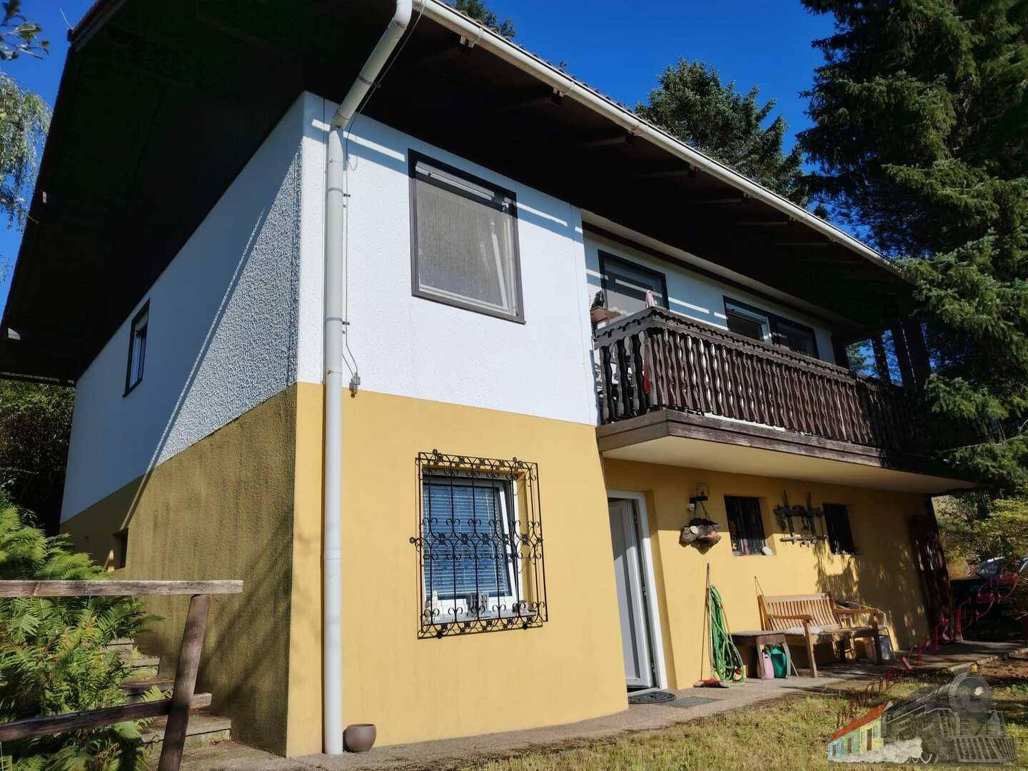 Ansicht des Hauses mit gelb-weißer Fassade, Holzbalkon und umgebender Natur.
