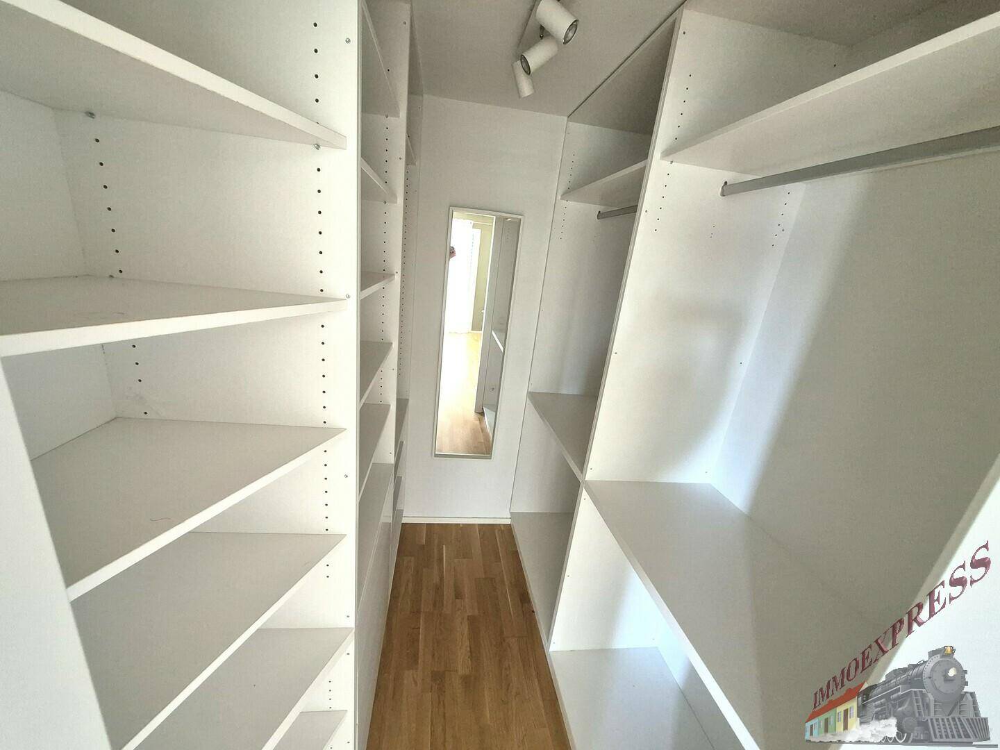 Begehbarer Kleiderschrank mit weißen Regalen und Holzfußboden, gut beleuchtet und geräumig.