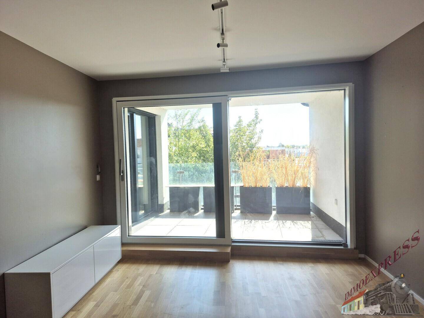 Helles Wohnzimmer mit Holzparkettboden und großem Schiebefenster zum Balkon mit Pflanzen.