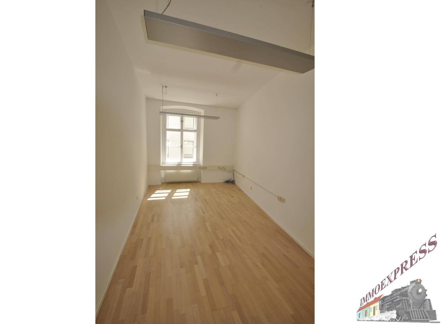 Schmaler Büroraum mit Holzboden, einem Fenster und einer modernen Deckenleuchte.
