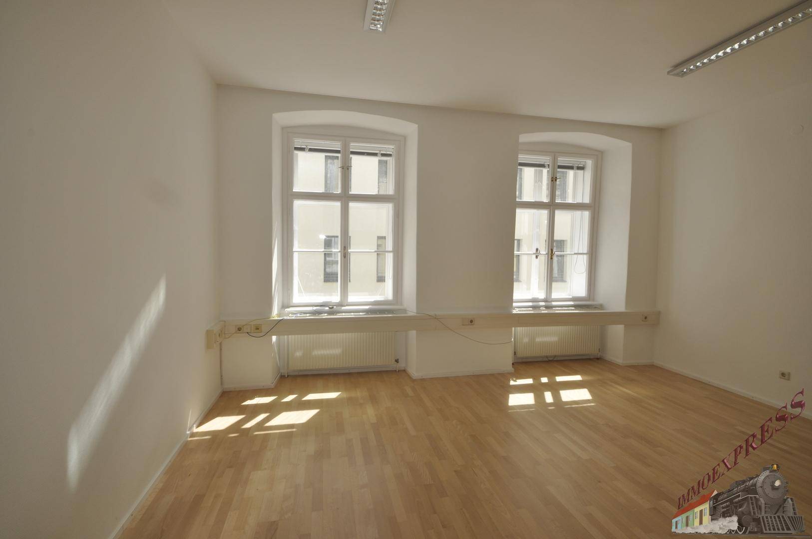 Leerer Büroraum mit Holzboden, weißen Wänden und zwei großen Fenstern.