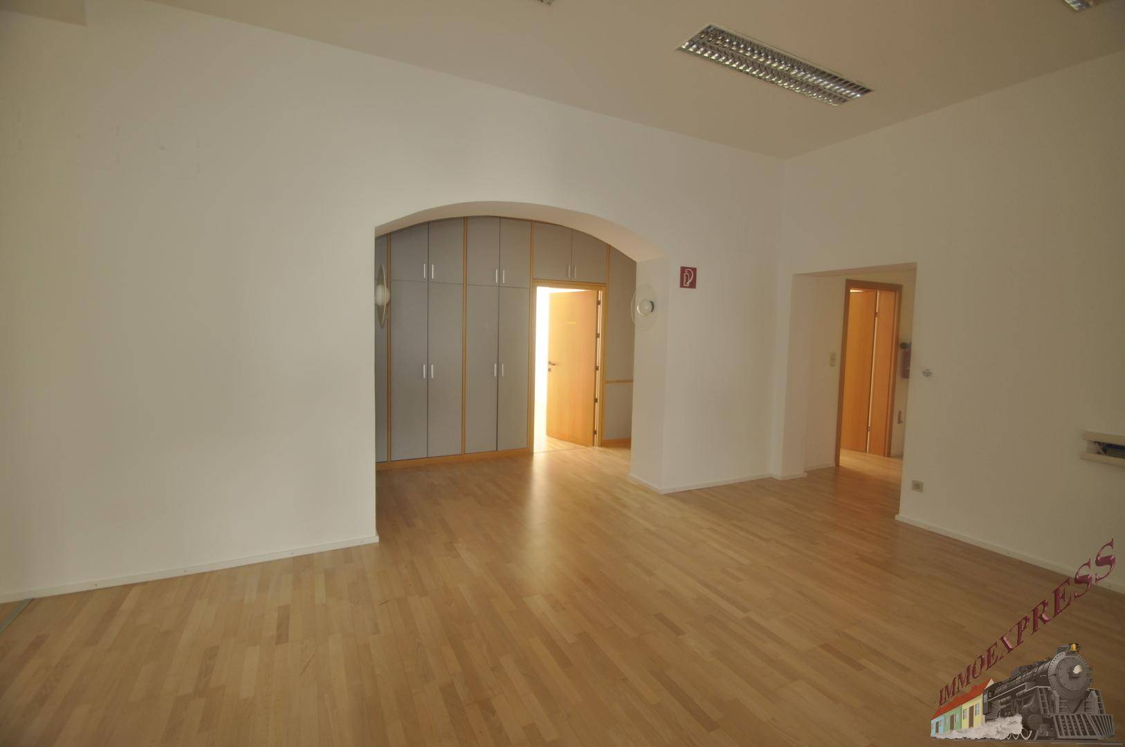 Büroraum mit Holzboden, Einbauschränken in einer Nische und Zugang zu weiteren Bereichen.