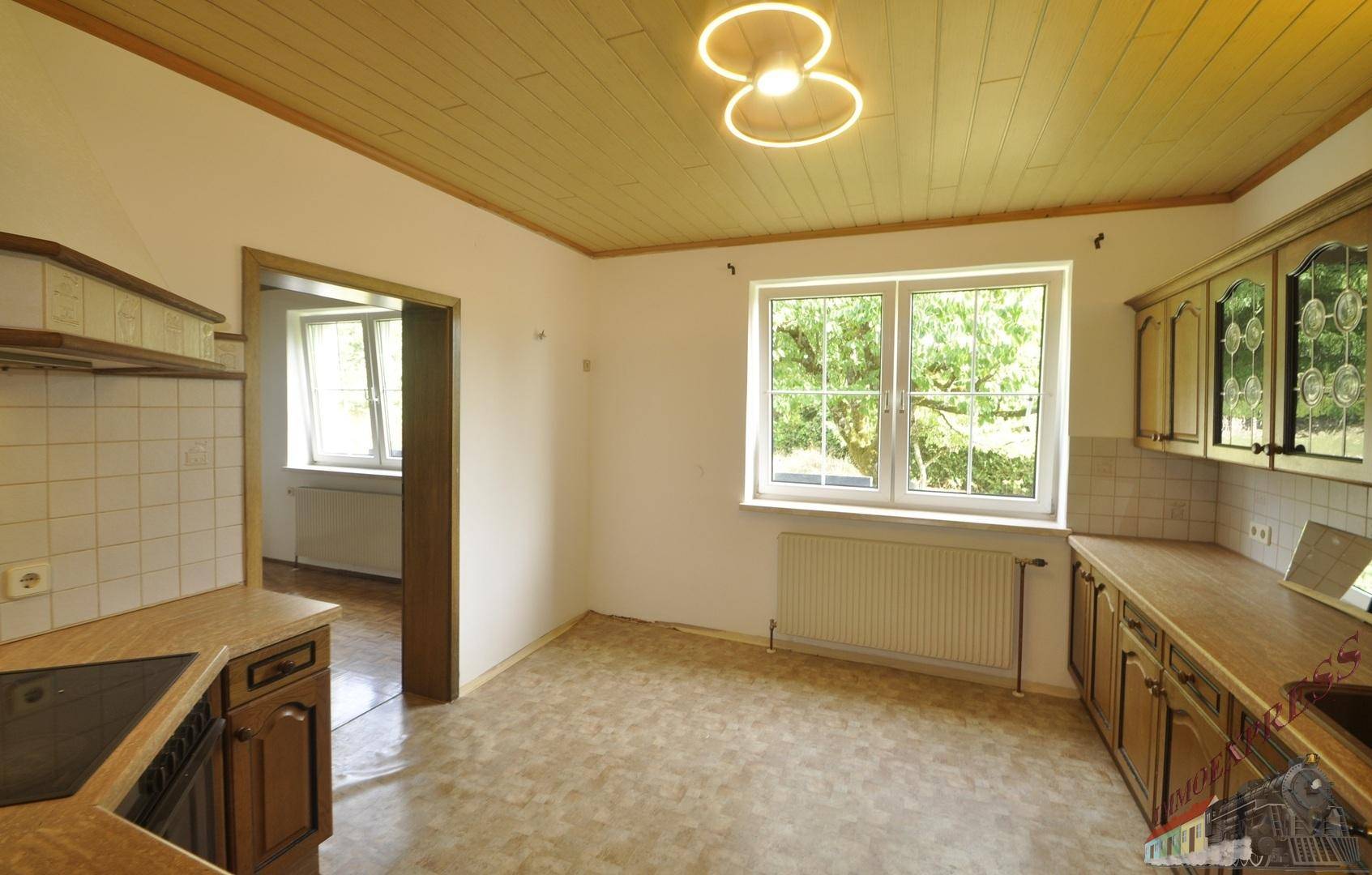 Helle Küche mit Holzschränken, Fliesenboden und einem Fenster mit Ausblick ins Grüne.