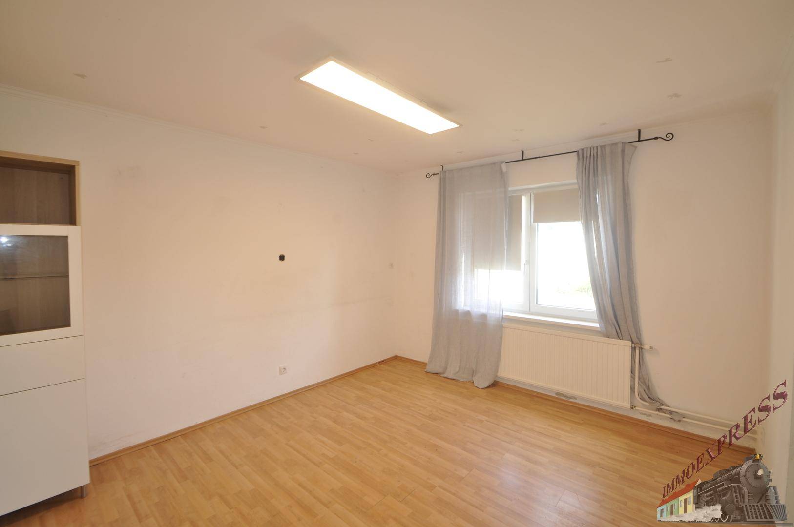 Helles Wohnzimmer mit Laminatboden, weißen Wänden und einem Fenster mit Vorhängen.