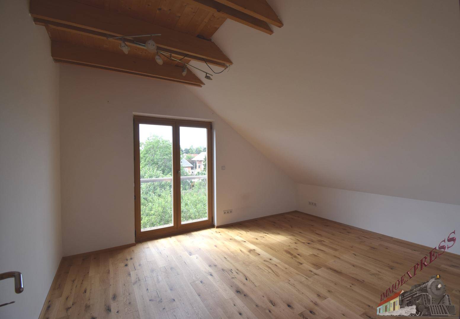 Lichtdurchflutetes Dachzimmer mit Holzboden und Zugang zu einem französischen Balkon mit grüner Aussicht.
