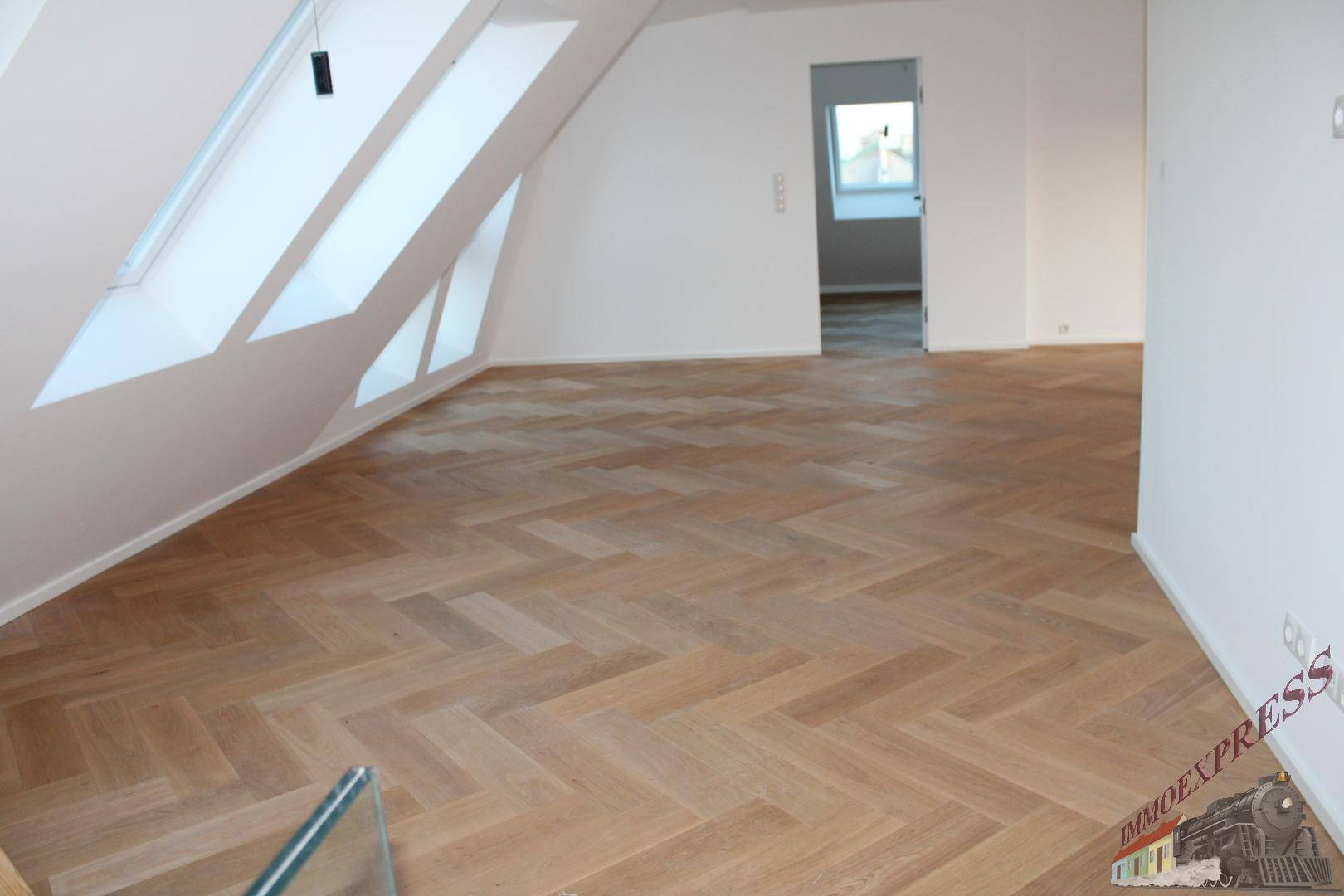 Geräumiges Dachgeschosszimmer mit hellem Fischgrätparkett und mehreren Dachfenstern.