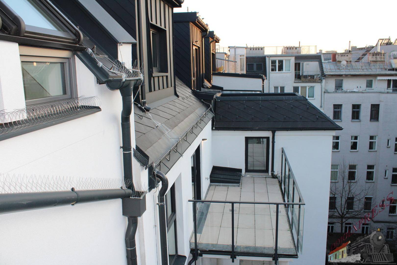 Kleiner Balkon mit Glasgeländer und Blick auf die umliegenden Dächer und Gebäude.