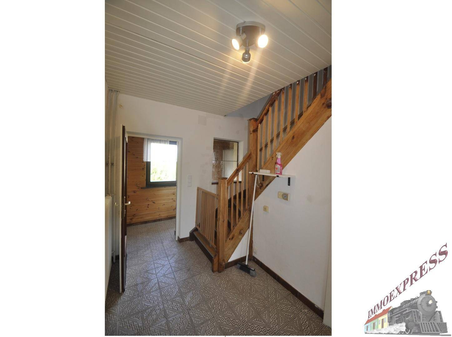 Heller Flur mit Holztreppe, gemusterten Fliesen und weißer Deckenverkleidung, die zum Obergeschoss führt.