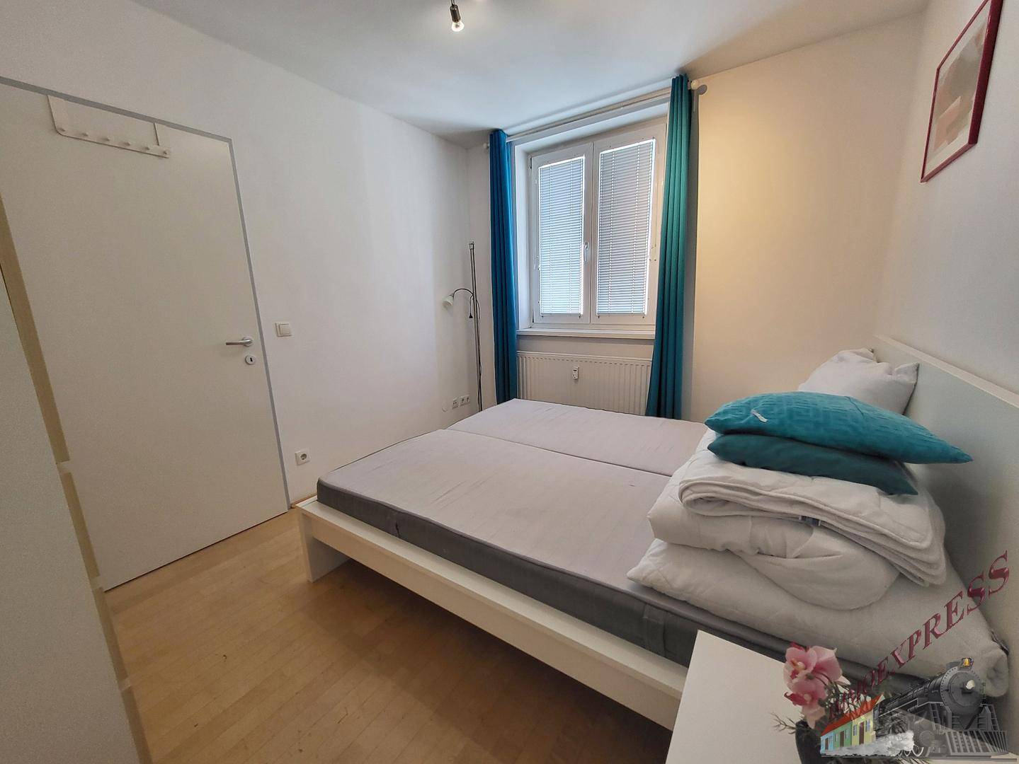 Helles Schlafzimmer mit Doppelbett, Holzfußboden und Fenster mit Jalousien.