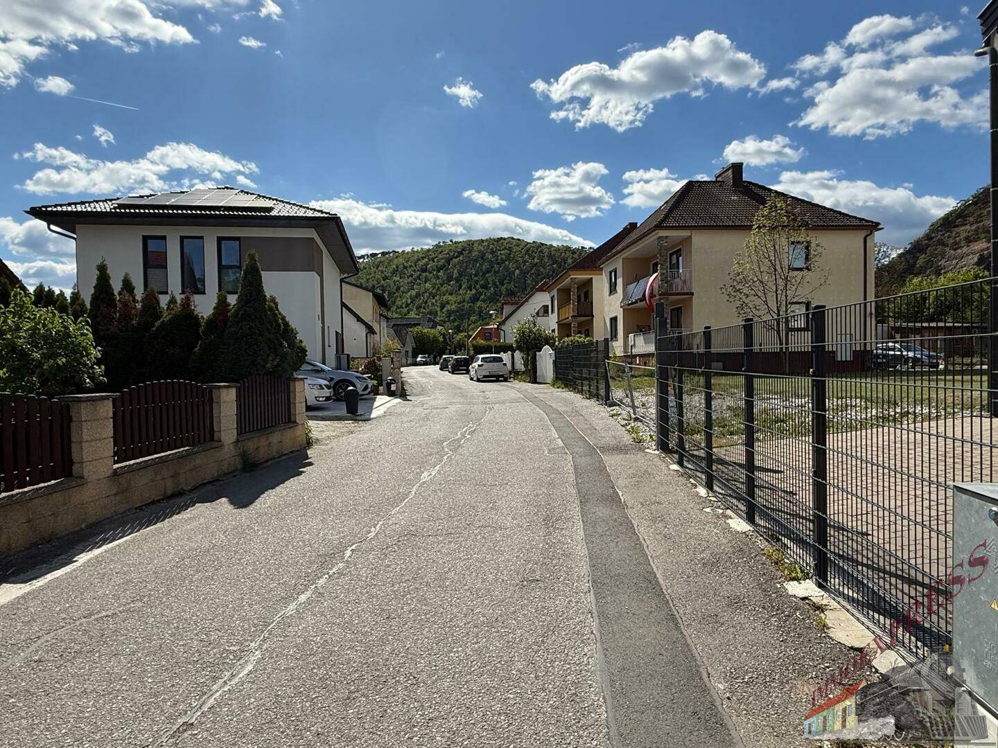 Wohnstraße mit Häusern auf beiden Seiten und geparkten Autos unter blauem Himmel.