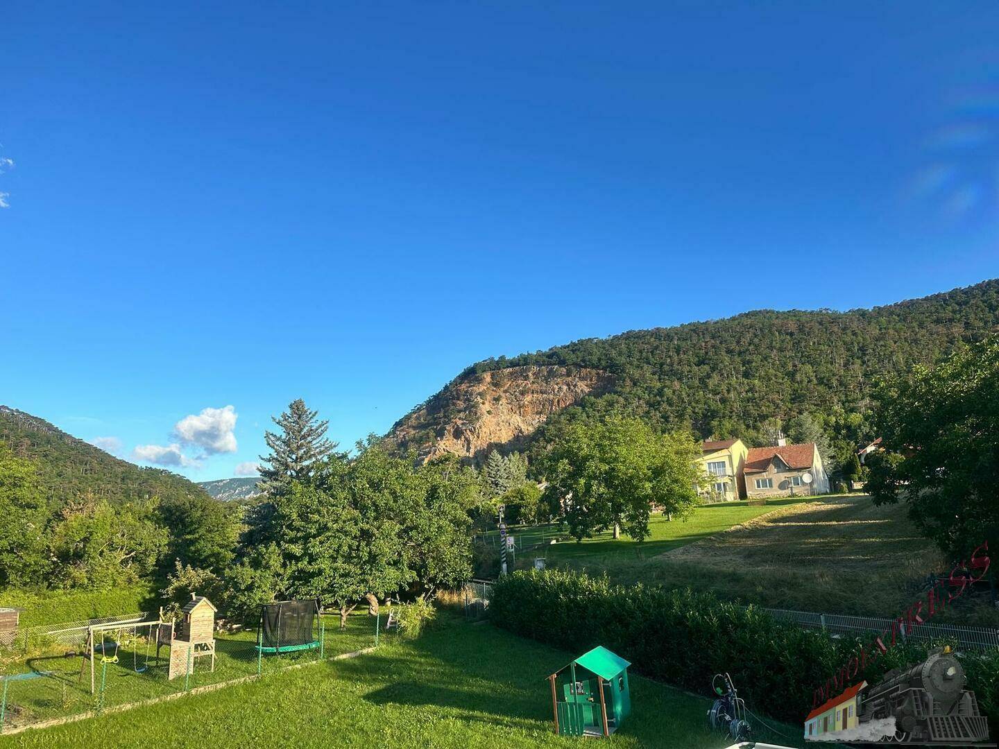 Weitläufiger Garten mit Spielplatz, Trampolin und Hecken, umgeben von grünen Hügeln und Bäumen.