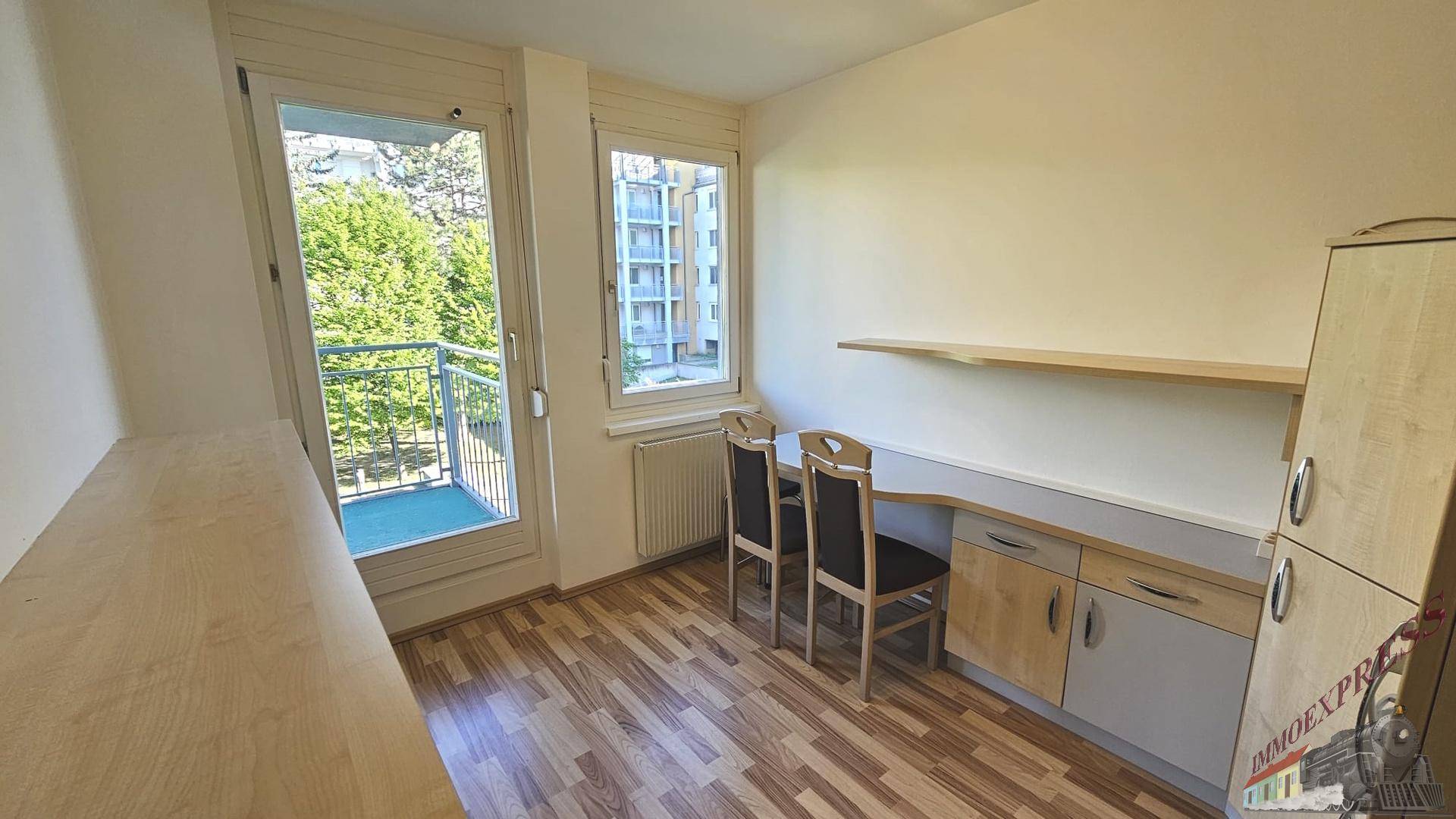 Helles Zimmer mit Schreibtisch, Regal und Zugang zu einem kleinen Balkon.