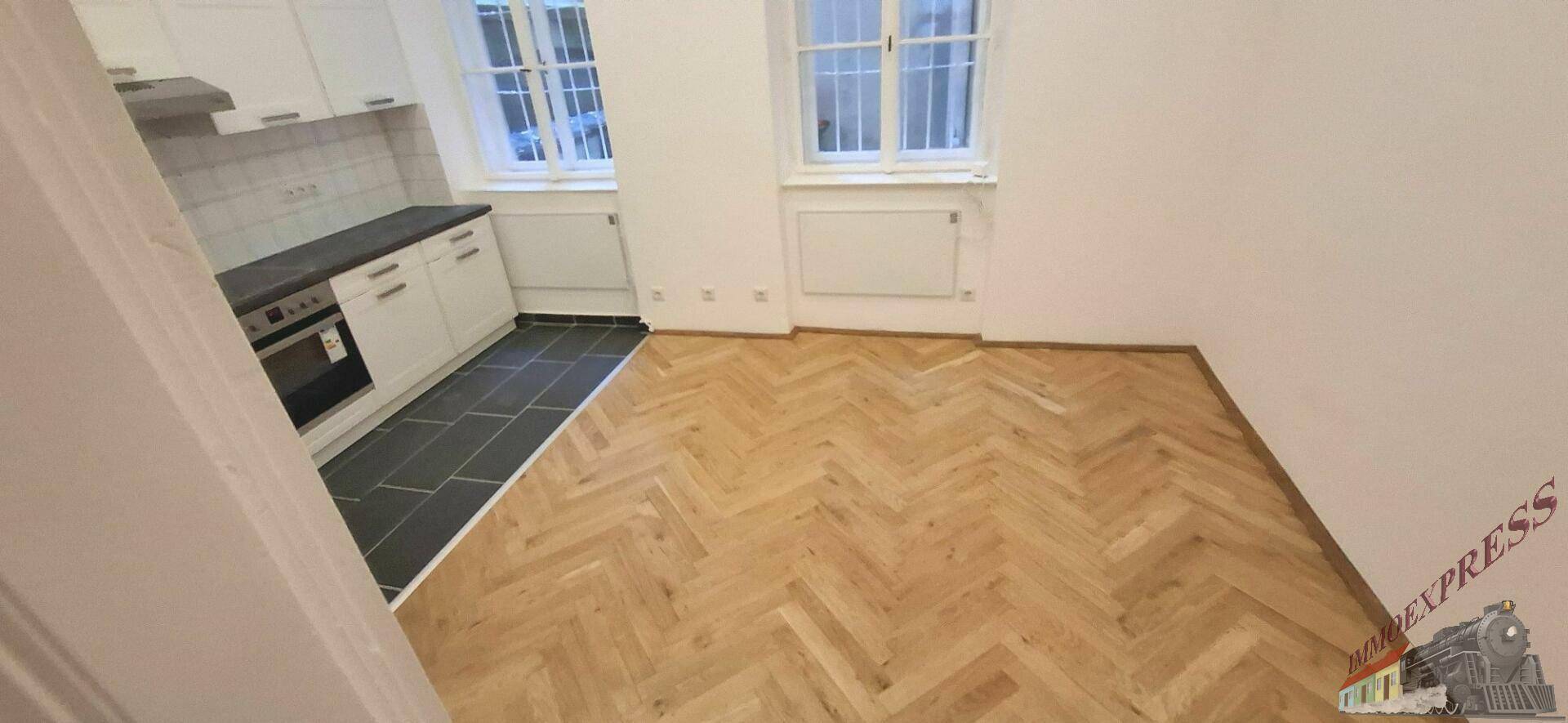 Offener Wohnbereich mit elegantem Fischgrätparkett und integrierter, funktionaler Küchenzeile am Fenster.