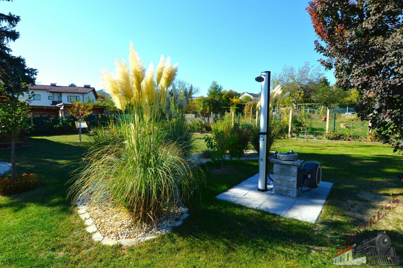 Gepflegter Gartenbereich mit Rasenfläche, Zierpflanzen und einer Außendusche für heiße Tage.